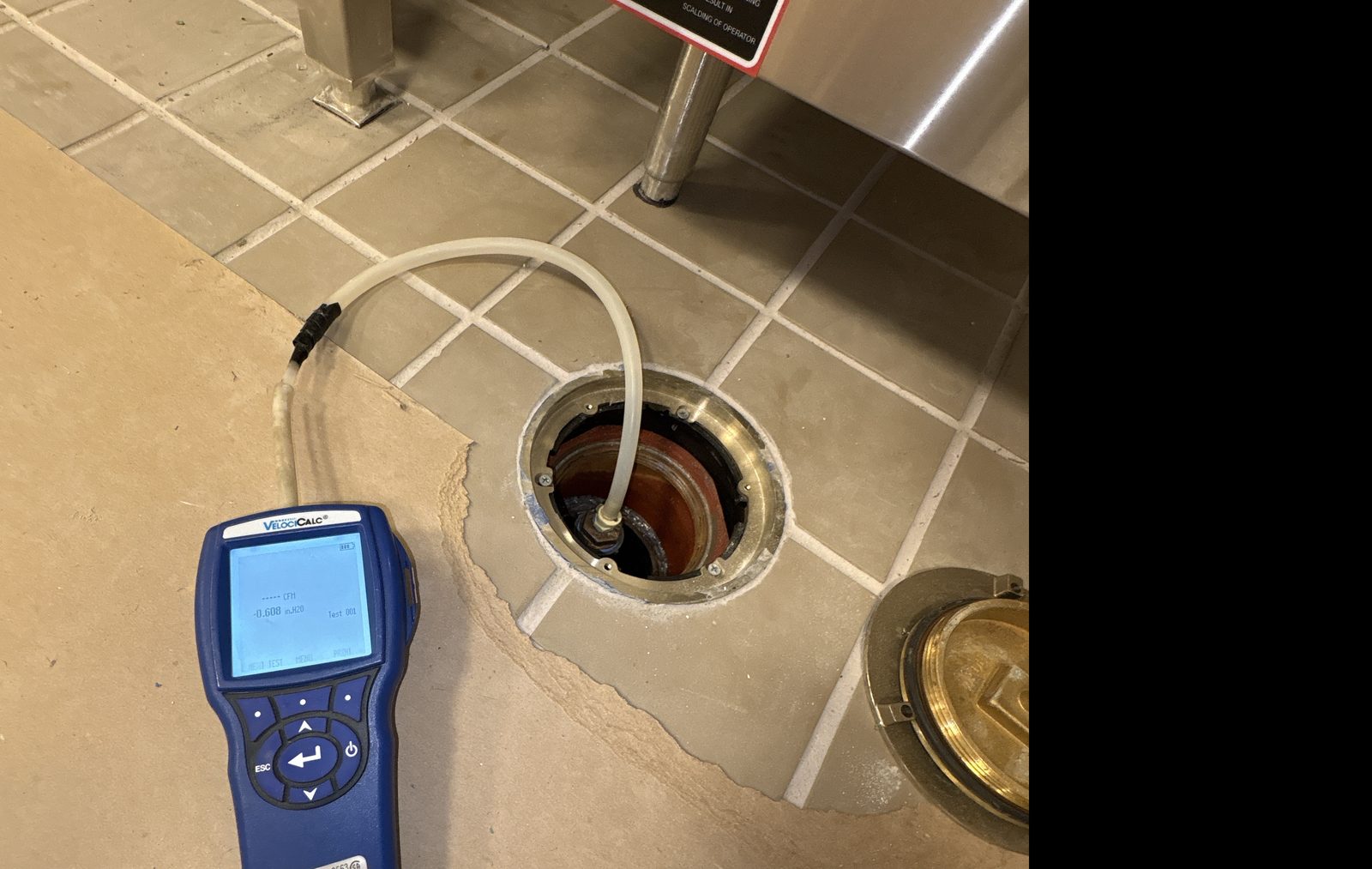 Digital micromanometer displaying a negative pressure reading connected by tubing to a floor-mounted sub-slab monitoring point during a sub-slab depressurization system inspection